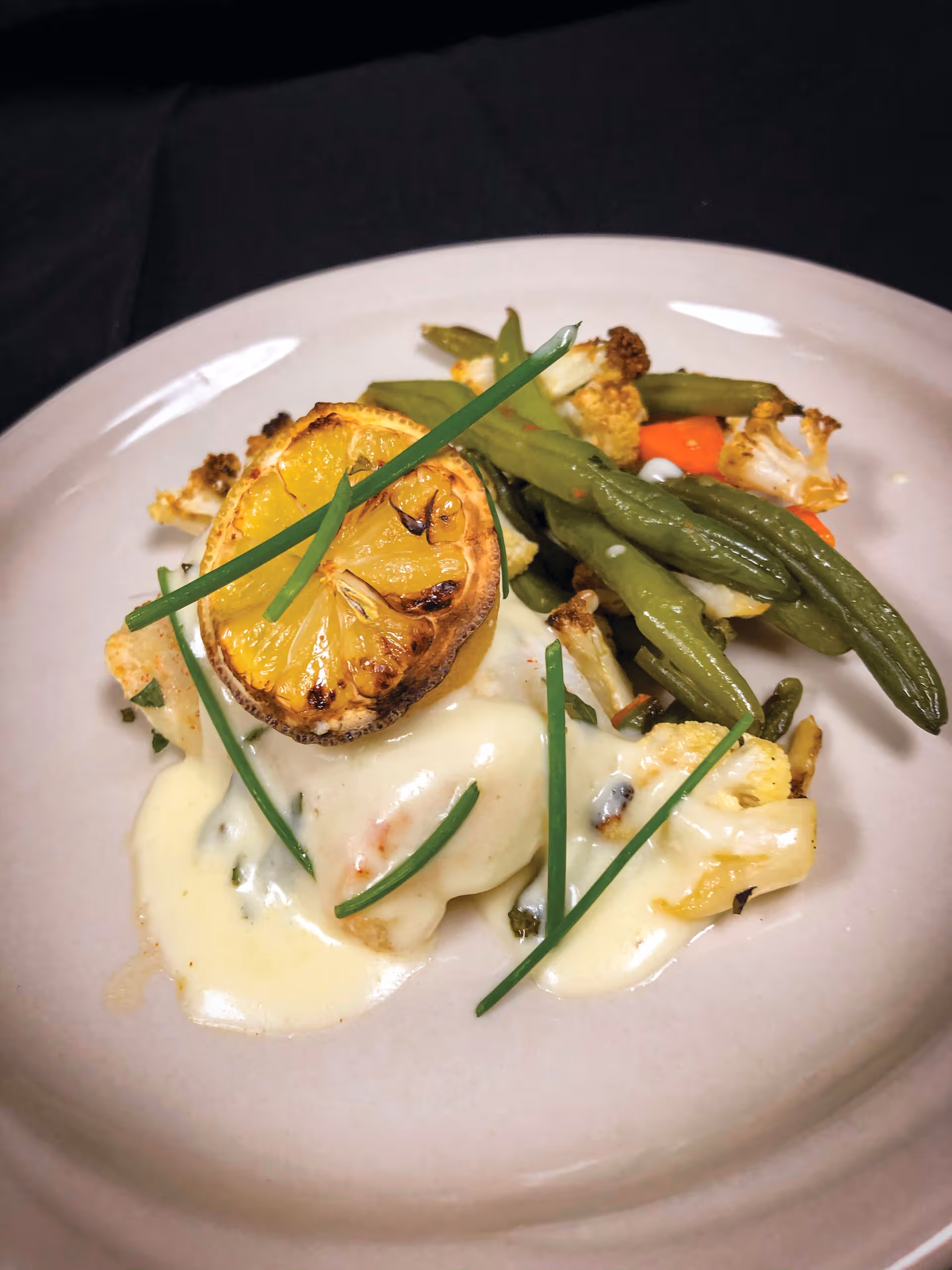 A plate of food featuring roasted green beans, cauliflower, and carrots topped with a creamy white sauce, garnished with a grilled lemon slice and fresh chives.