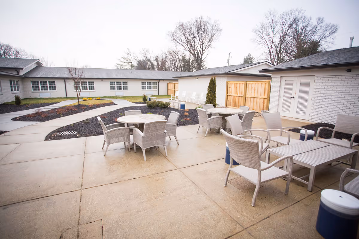 Outdoor patio area at Dominion Senior Living of Louisville featuring multiple round and rectangular tables with wicker chairs arranged on a concrete surface. The patio is surrounded by low garden beds with mulch and small shrubs, and the background shows single-story white brick buildings with windows and a wooden fence. Leafless trees are visible beyond the buildings under an overcast sky.