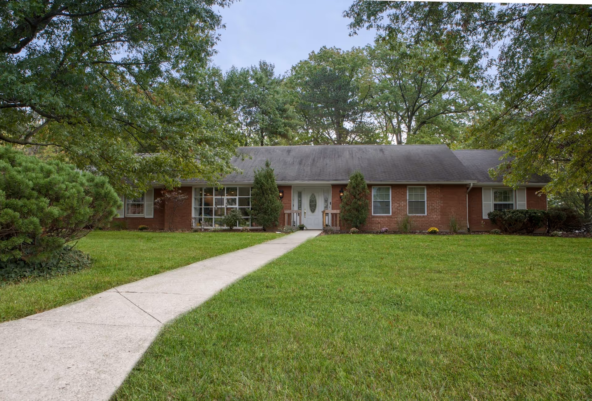 A single-story brick house with a gray shingled roof, white front door with glass panel, and several windows. The house is surrounded by green grass, trees, and shrubs, with a concrete walkway leading up to the entrance.