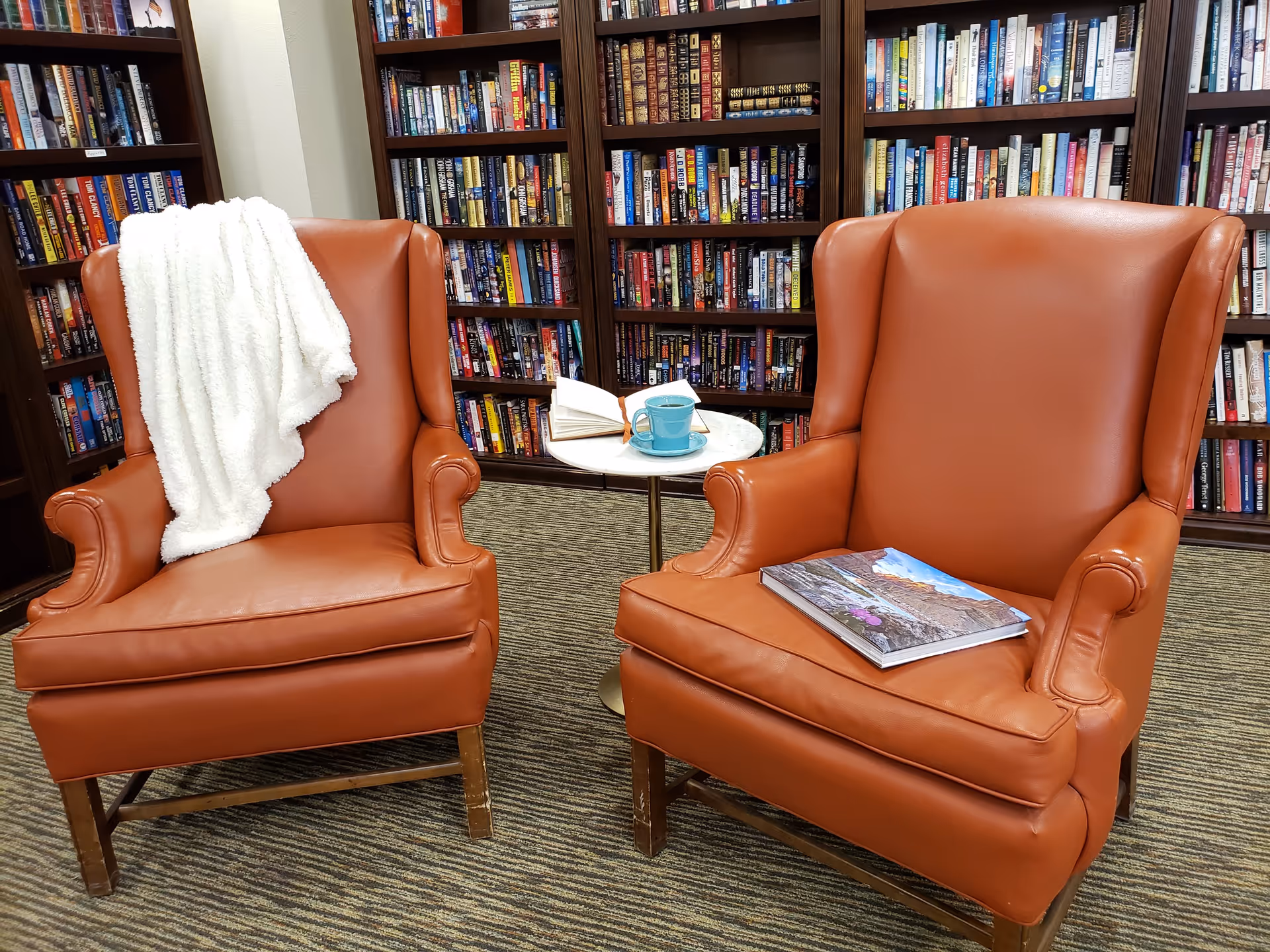 Two brown leather armchairs in front of bookshelves filled with books. One chair has a white blanket draped over the back, and the other has a book placed on the seat. Between the chairs is a small round table with an open book and a blue cup and saucer. The setting appears to be a cozy reading area in a library or common room.