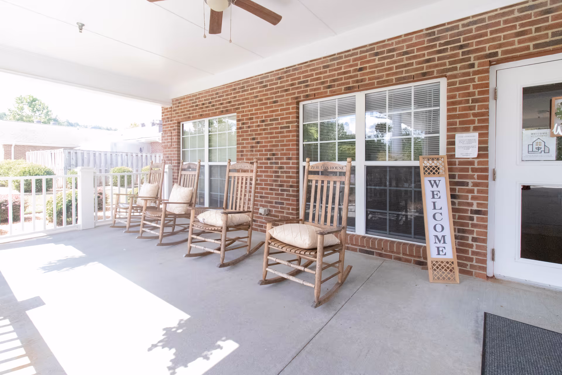 Covered outdoor porch area with four wooden rocking chairs, each with a cushion, against a brick wall with windows. A vertical sign reading 'WELCOME' leans against the wall near a white door. The porch has a ceiling fan and overlooks a garden area with bushes and a wooden fence.