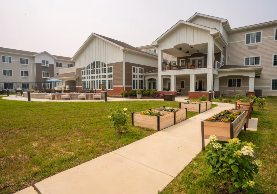 Exterior view of a multi-story senior living building with a covered patio, outdoor seating, raised garden beds, and a walkway.