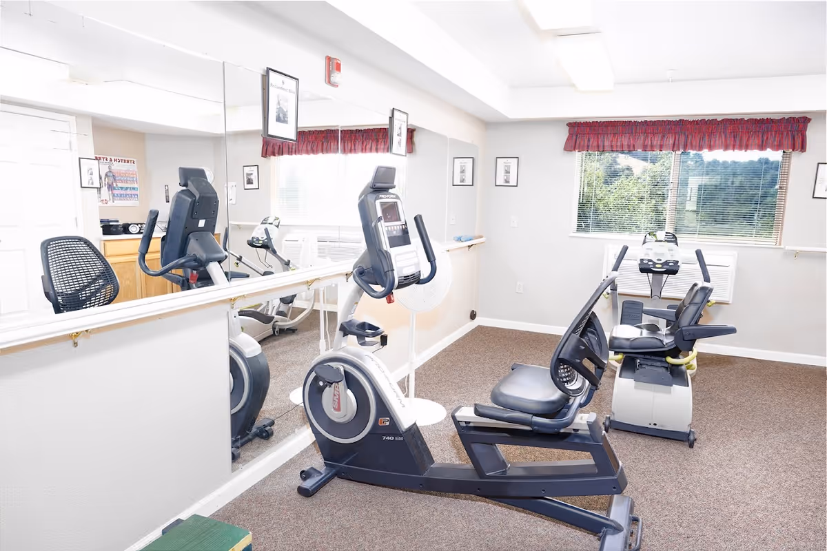 A small exercise room with two recumbent stationary bikes facing a large wall mirror. The room has beige walls, a window with a red valance, and carpeted flooring. There are framed pictures on the walls and a white fan positioned between the bikes.