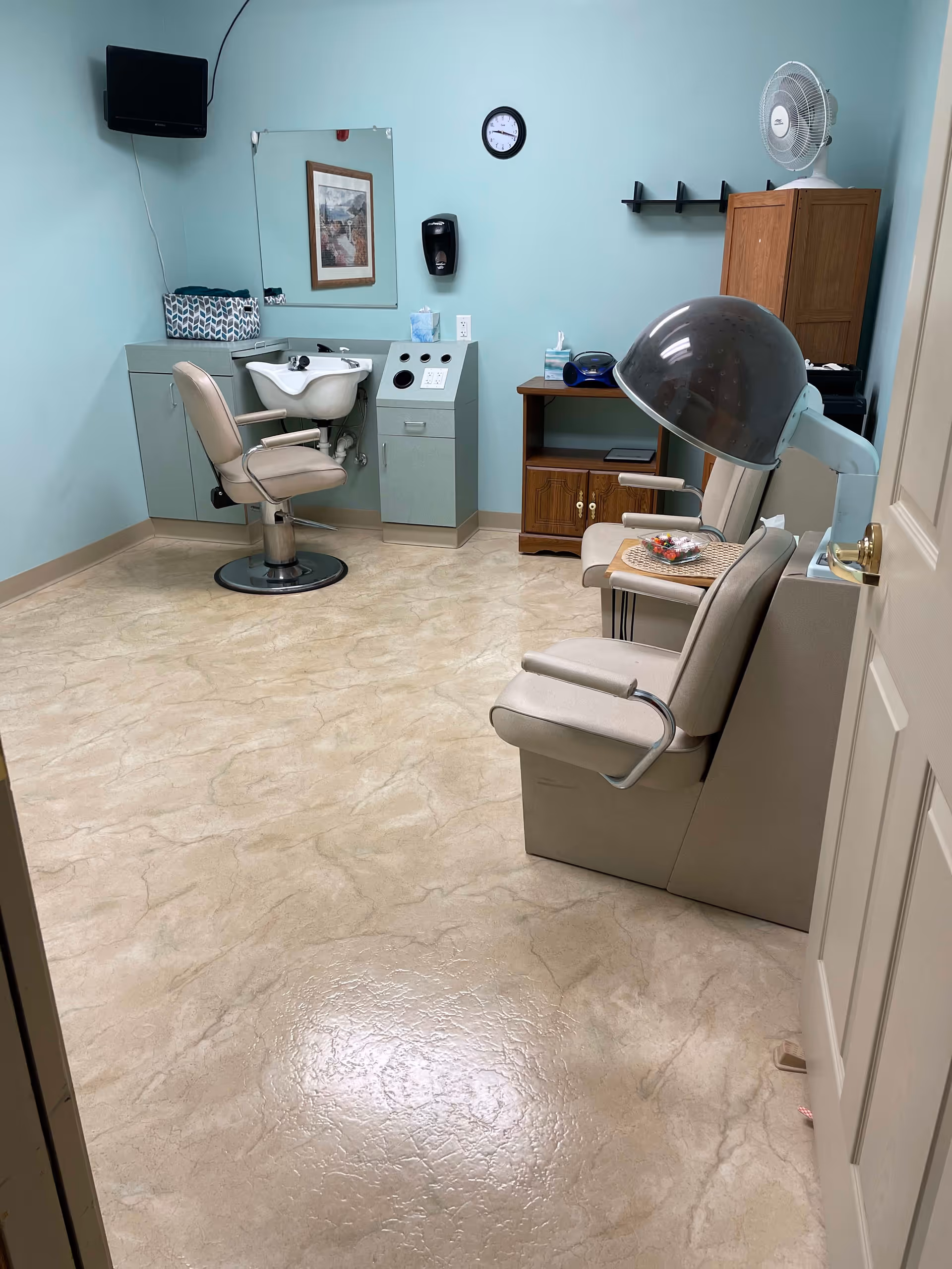 A small salon room with a hair washing station including a chair and sink, a hair drying chair with a hooded dryer, a small wooden cabinet with a fan on top, a wall-mounted TV, a clock, and a mirror. The walls are painted light blue and the floor has a beige marble-like pattern.