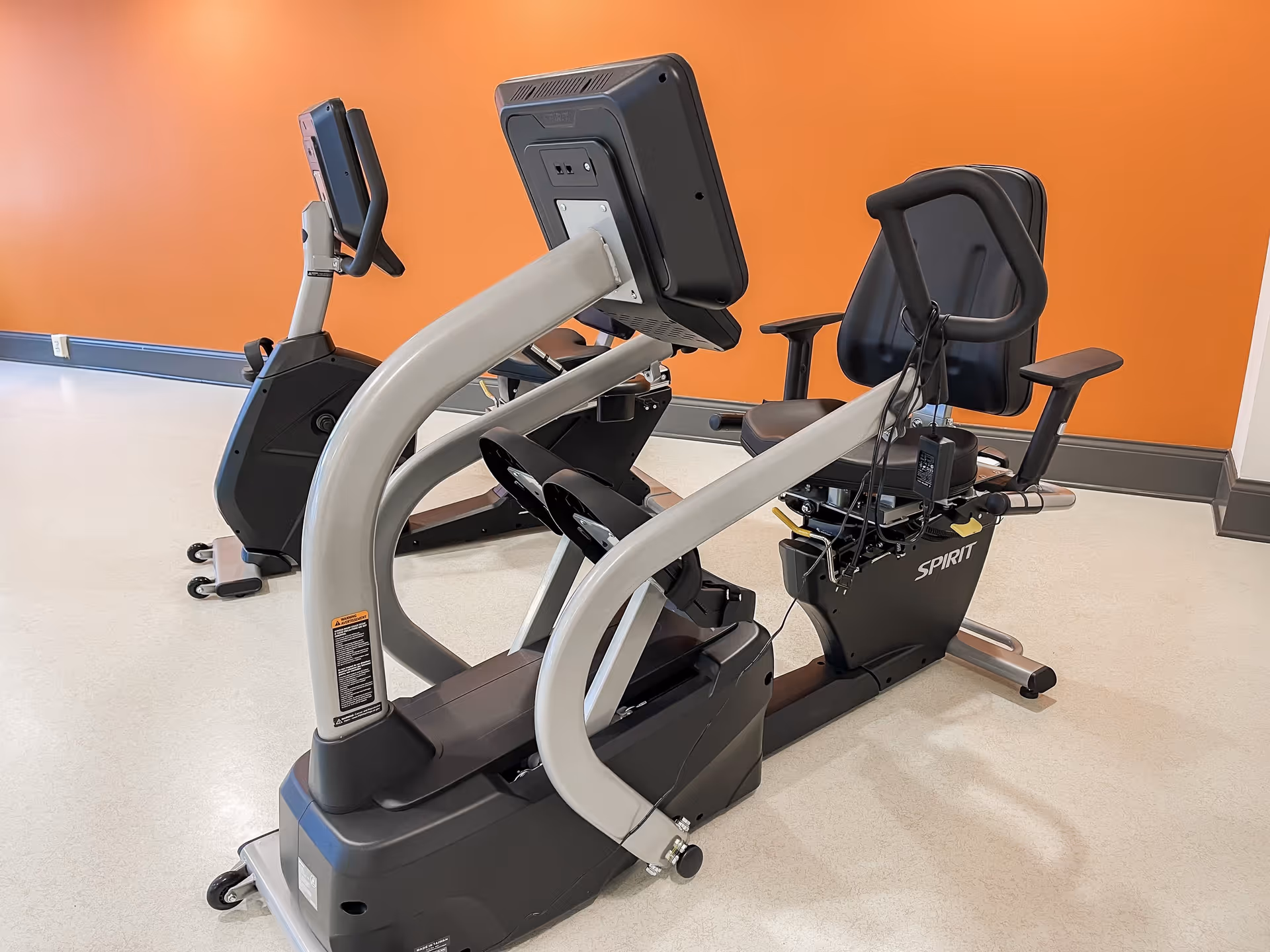 Two stationary recumbent exercise bikes with digital screens in a fitness room with an orange wall and light-colored flooring.