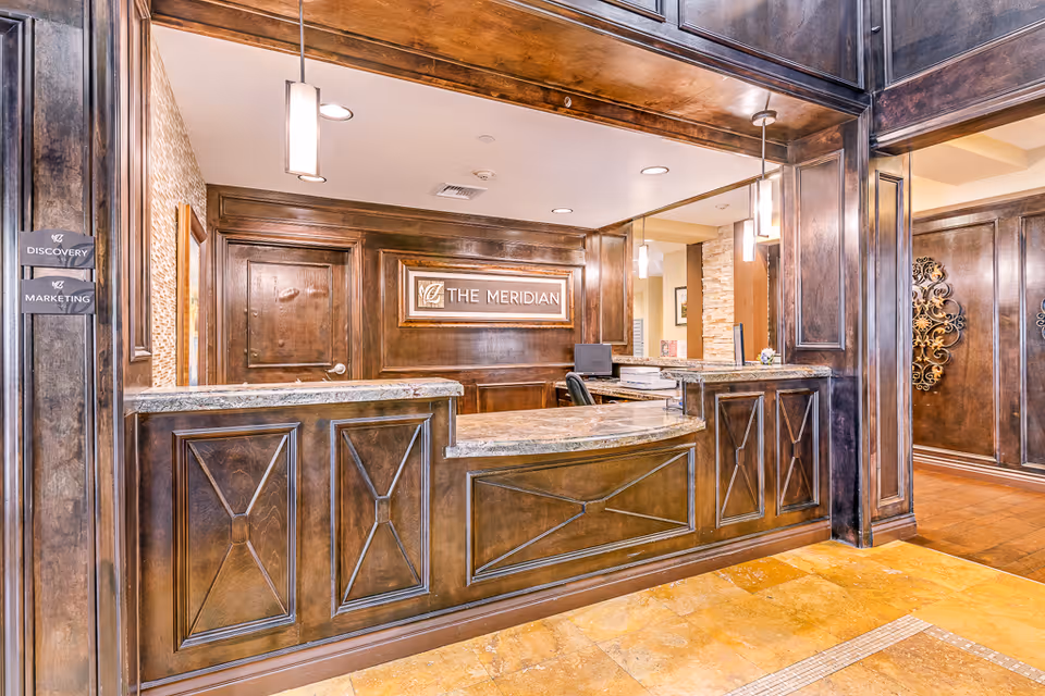 Reception desk area with dark wood paneling and marble countertop. Behind the desk is a sign that reads 'THE MERIDIAN'. Two pendant lights hang from the ceiling above the desk. There are signs on the left wall indicating directions to 'DISCOVERY' and 'MARKETING'. The floor is tiled with a warm yellowish tone.