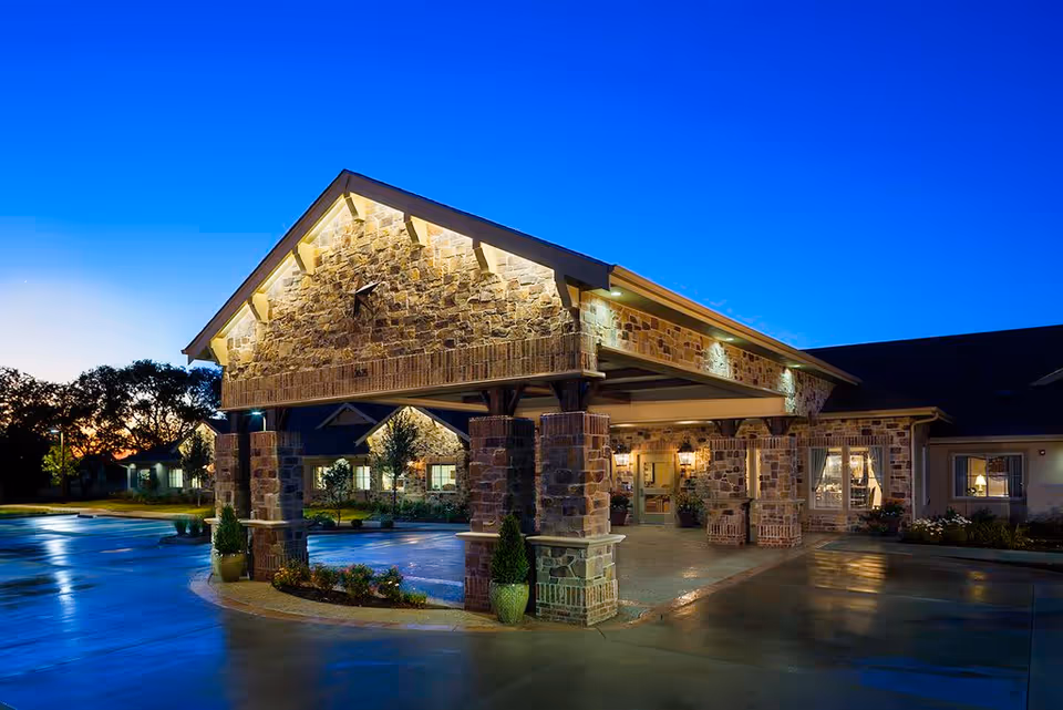 Exterior view of a stone and brick building entrance at dusk with warm lighting under a covered driveway and surrounding landscaping.