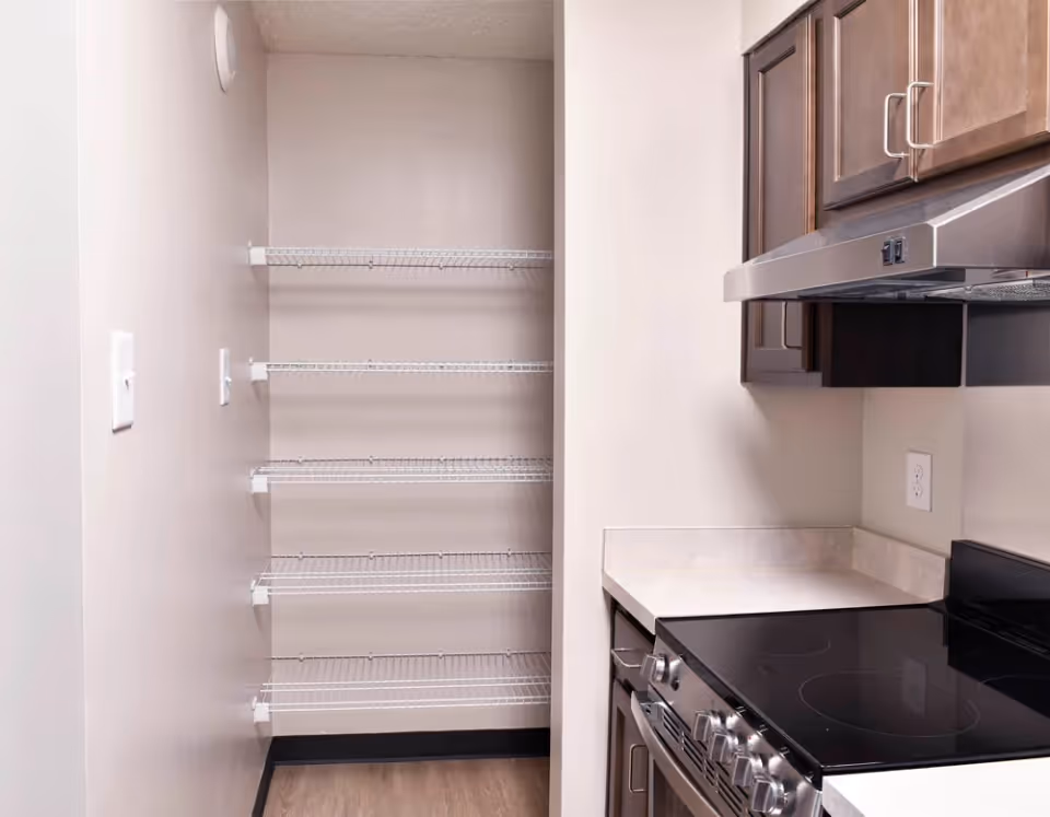 A small kitchen area featuring a walk-in pantry with wire shelves beside a stove and dark wood cabinets.