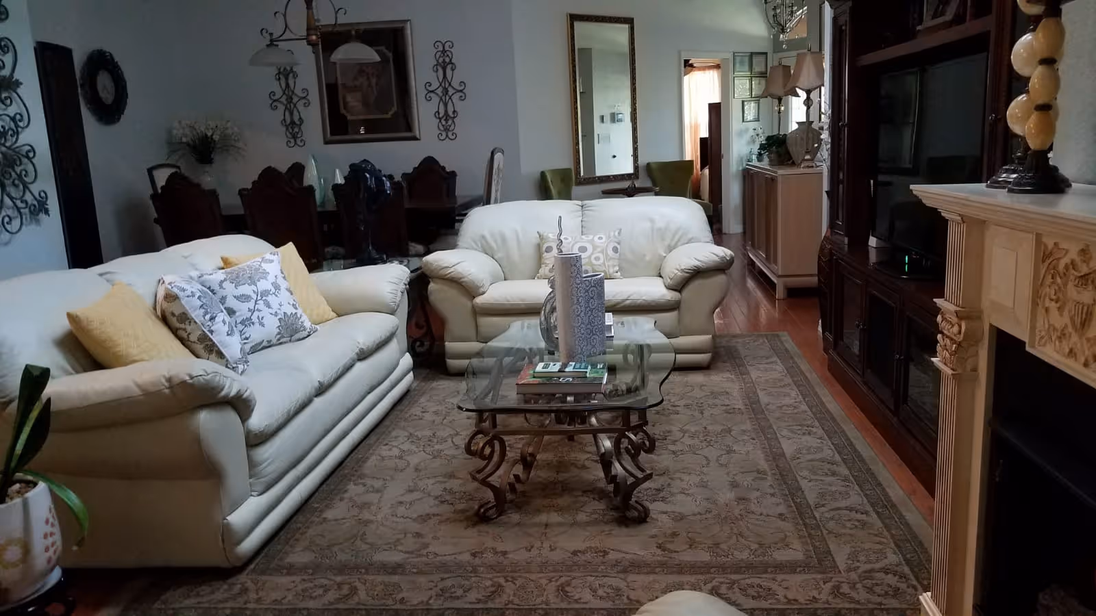 A cozy living room with two cream-colored sofas adorned with decorative pillows, a glass coffee table with books and vases, a patterned area rug, a wooden entertainment center with a TV, and a fireplace with ornate detailing. In the background, there is a dining area with a dark wooden table and chairs, wall decorations, and a large mirror reflecting part of the room.