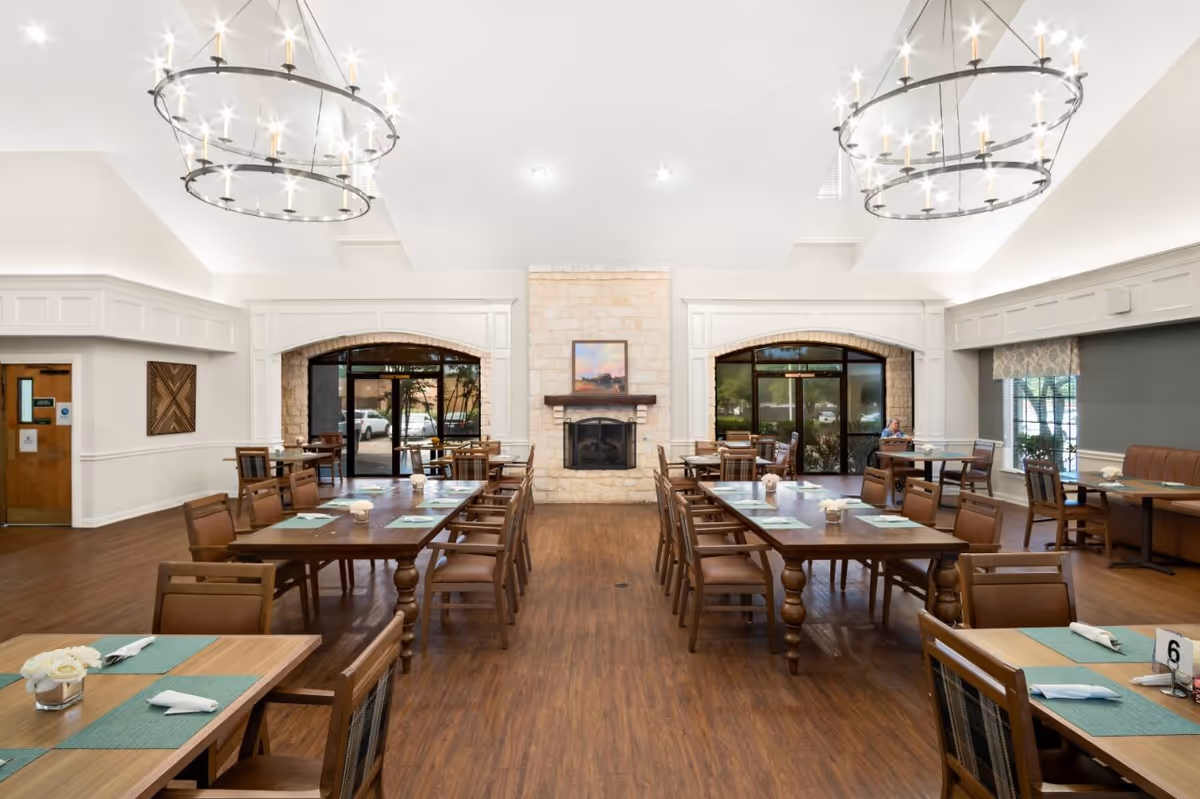 Spacious dining room with wooden tables and chairs arranged neatly. The room features two large circular chandeliers hanging from a high white ceiling, a stone fireplace with a painting above it, and large windows and doors letting in natural light. The floor is wooden, and the tables are set with placemats, napkins, and small flower arrangements.