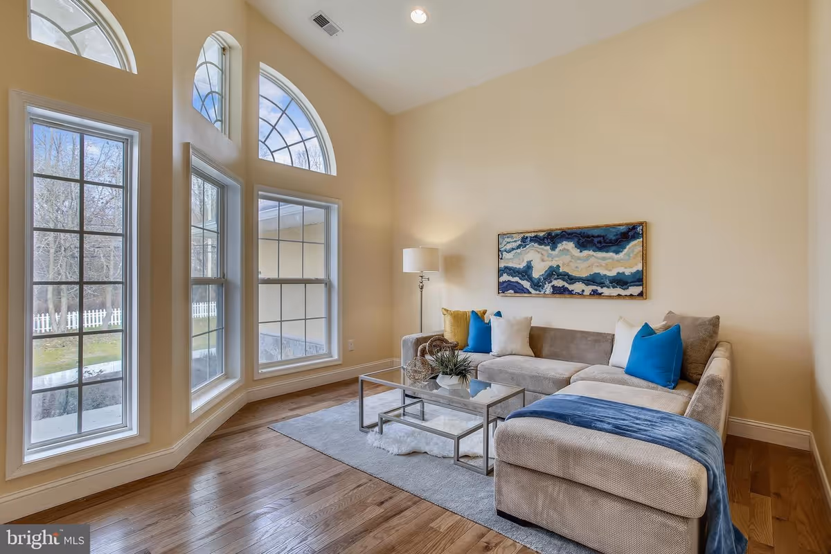 Bright living room with large arched windows, a beige sectional sofa with blue pillows and throw, glass coffee table, and hardwood floors.