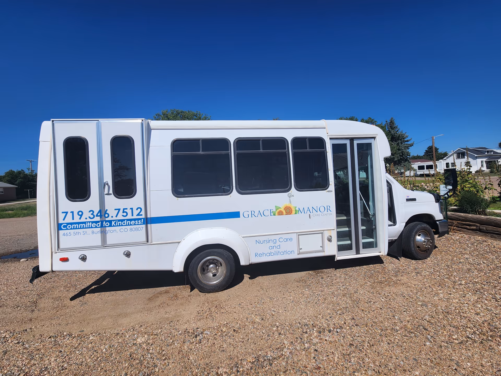 White Grace Manor Care Center shuttle bus parked on gravel with logo and phone number visible against a clear blue sky.