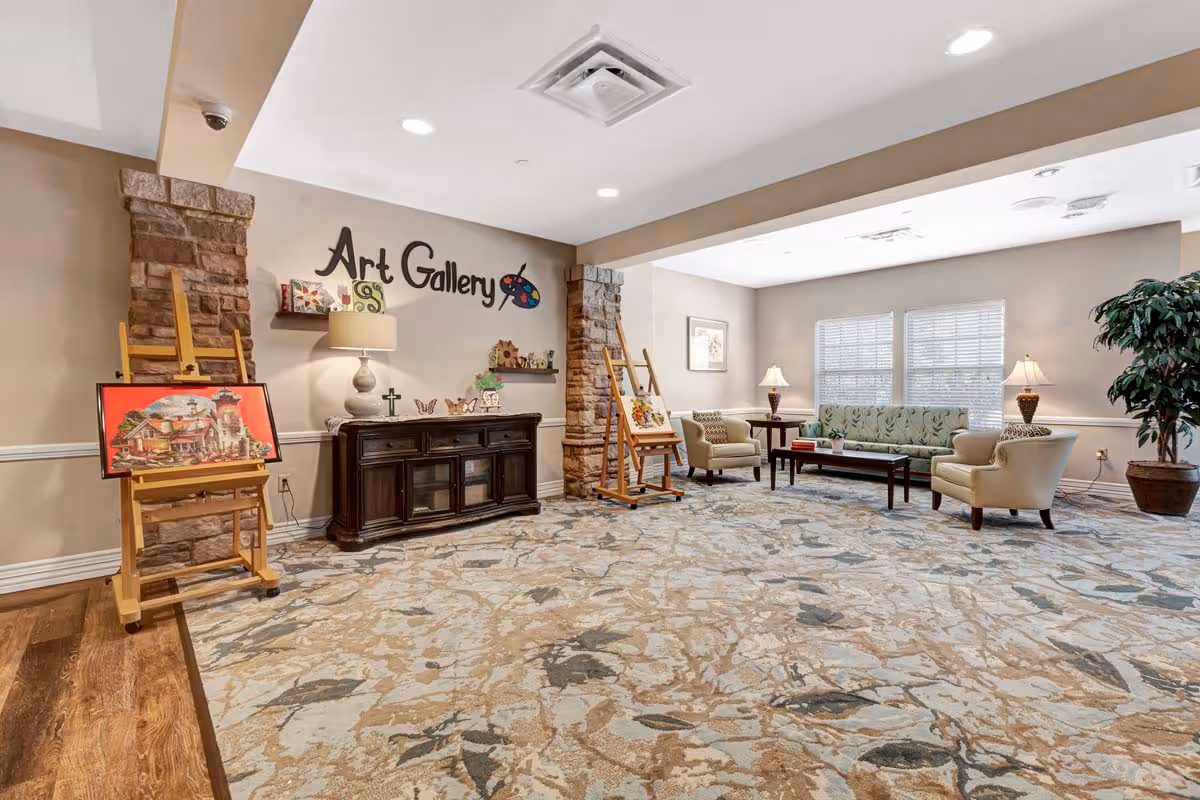 A spacious, well-lit art gallery room in a senior living facility featuring two wooden easels displaying paintings, a dark wooden sideboard with decorative items, a beige armchair, a green patterned sofa, two beige armchairs, a coffee table, two table lamps, a large potted plant, and a patterned carpet. The wall has the words 'Art Gallery' with a paint palette graphic.