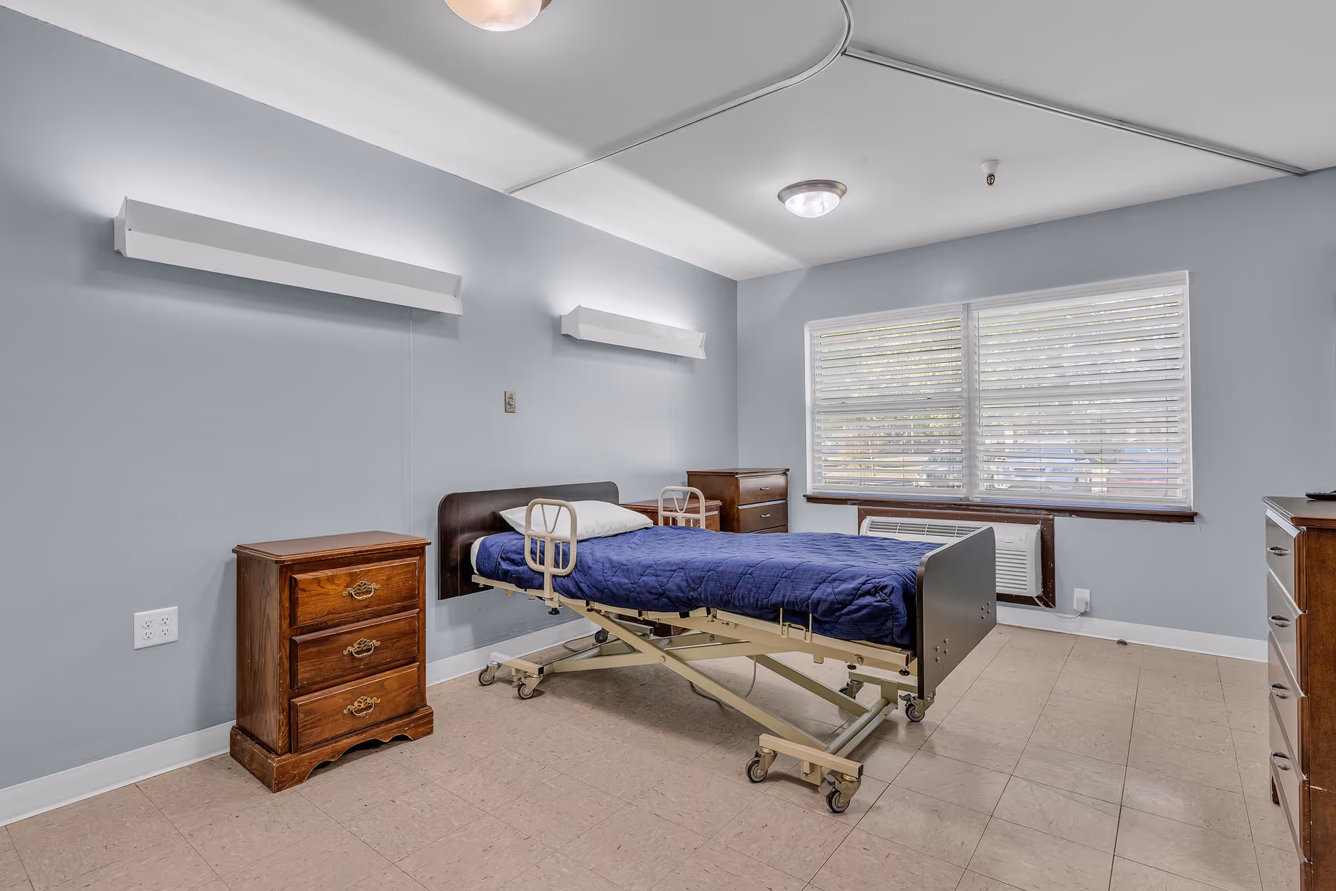 A simple healthcare facility bedroom with a hospital bed covered in a blue blanket, two wooden nightstands, a wooden dresser, and a large window with white blinds letting in natural light. The walls are painted light blue and the floor is tiled.
