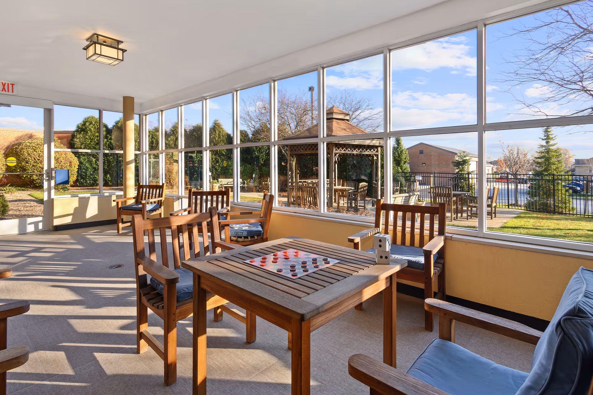 A sunlit indoor seating area with wooden chairs and tables, one table set up with a checkers game and dice. Large windows provide a clear view of an outdoor patio area with more wooden tables and chairs, greenery, and a gazebo under a blue sky.