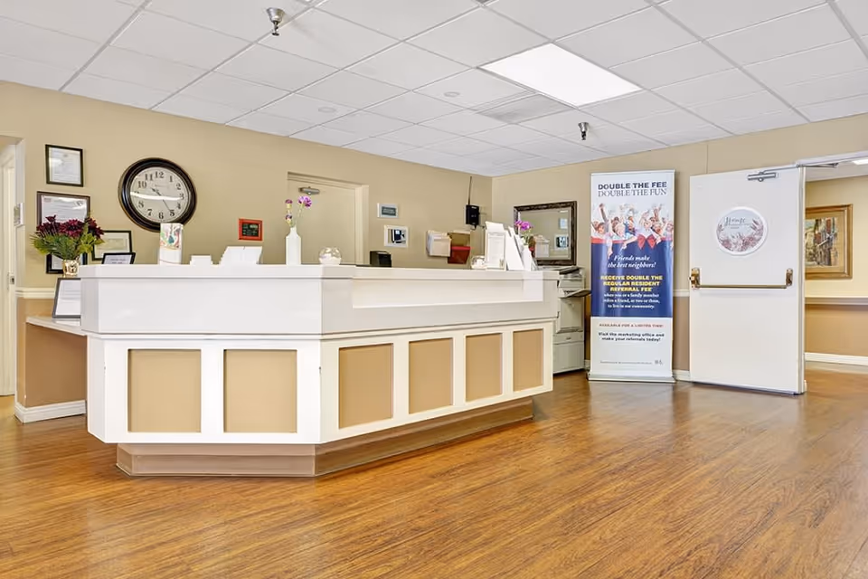 Reception area of Anaheim Villa senior living facility with a white and beige front desk, a wall clock, framed certificates, a flower vase, and a promotional banner near an open door. The floor is wooden and the walls are painted beige.