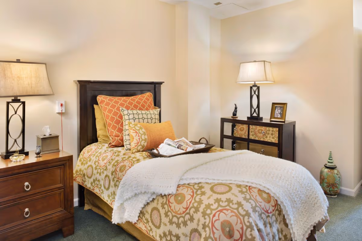 A cozy bedroom with a single bed featuring a patterned bedspread and multiple decorative pillows. There is a wooden nightstand on the left with a lamp, tissue box, and small decorative items. On the right side, there is a small dresser with a lamp, a framed photo, and a small figurine. A white knitted blanket is draped over the foot of the bed, and a decorative vase is placed on the floor in the corner.