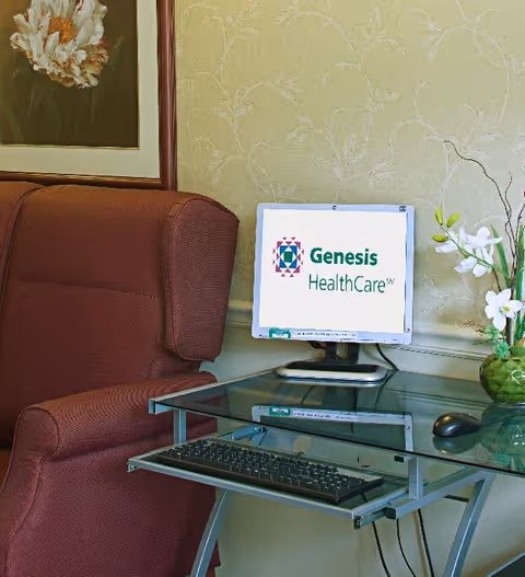 A cozy corner with a maroon upholstered armchair next to a glass computer desk. On the desk is a monitor displaying the Genesis HealthCare logo, a keyboard, and a mouse. A green vase with white flowers is also on the desk, and a framed floral painting hangs on the wall behind the chair.