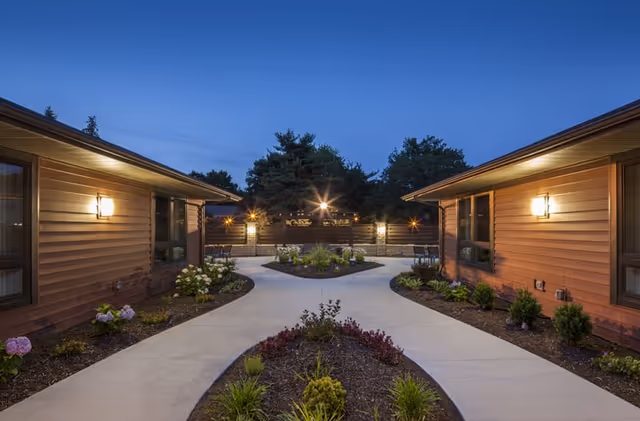 Evening view of a landscaped outdoor courtyard area between two single-story buildings with brown siding, illuminated wall lights, paved walkways, and various plants and shrubs.