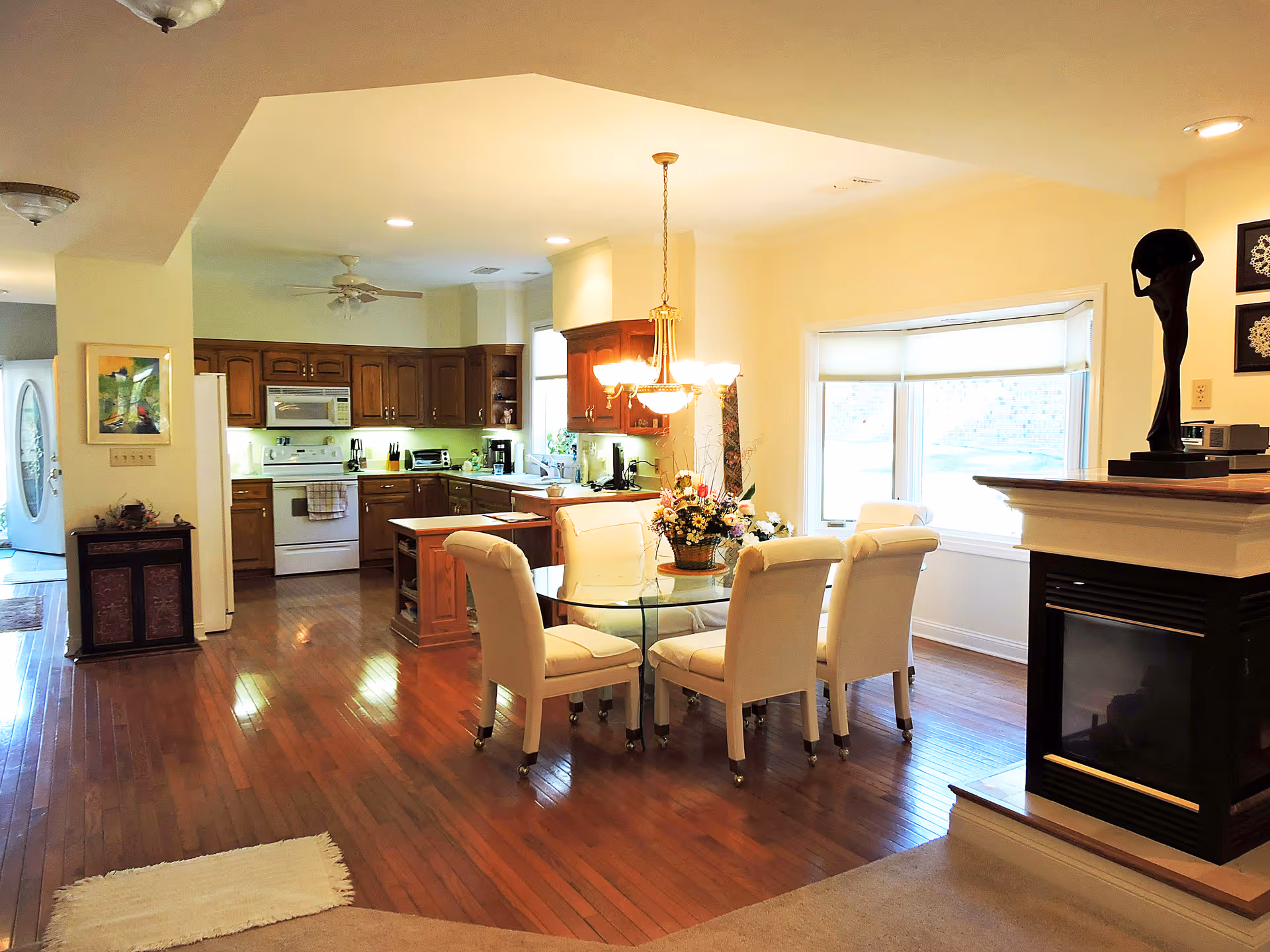 A bright and spacious open-plan kitchen and dining area with wooden floors. The kitchen has wooden cabinets, white appliances including a stove, microwave, and refrigerator. The dining area features a round glass table with six white upholstered chairs and a floral centerpiece. A large window with a white blind lets in natural light. To the right, there is a fireplace with a black sculpture on the mantel. The walls are painted a soft yellow, and the ceiling has recessed lighting and a hanging chandelier above the dining table.