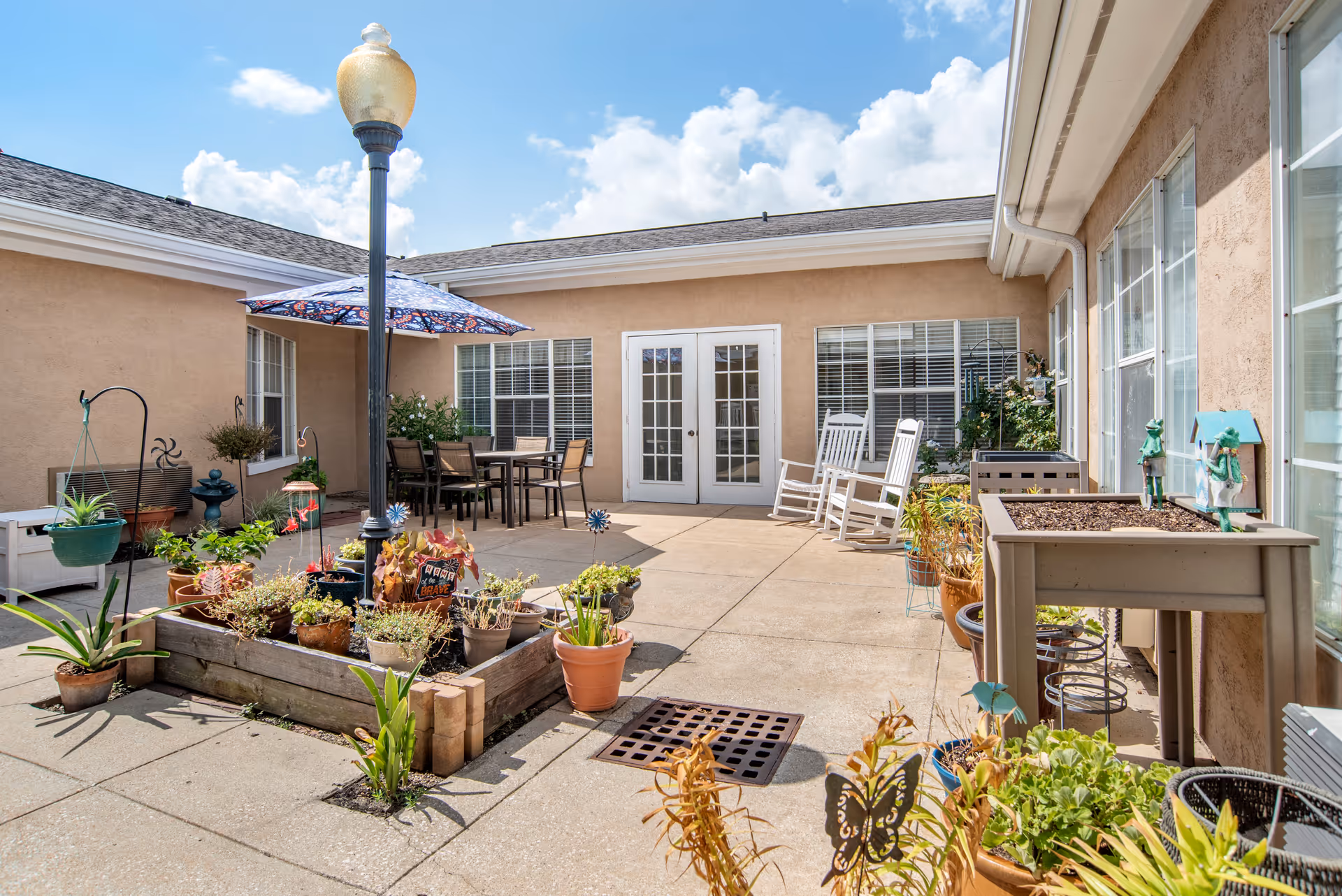 Outdoor patio area with potted plants, a raised garden bed, a table with chairs under a colorful umbrella, two white rocking chairs, and a lamppost. The patio is surrounded by beige walls with windows and a set of white double doors under a blue sky with some clouds.