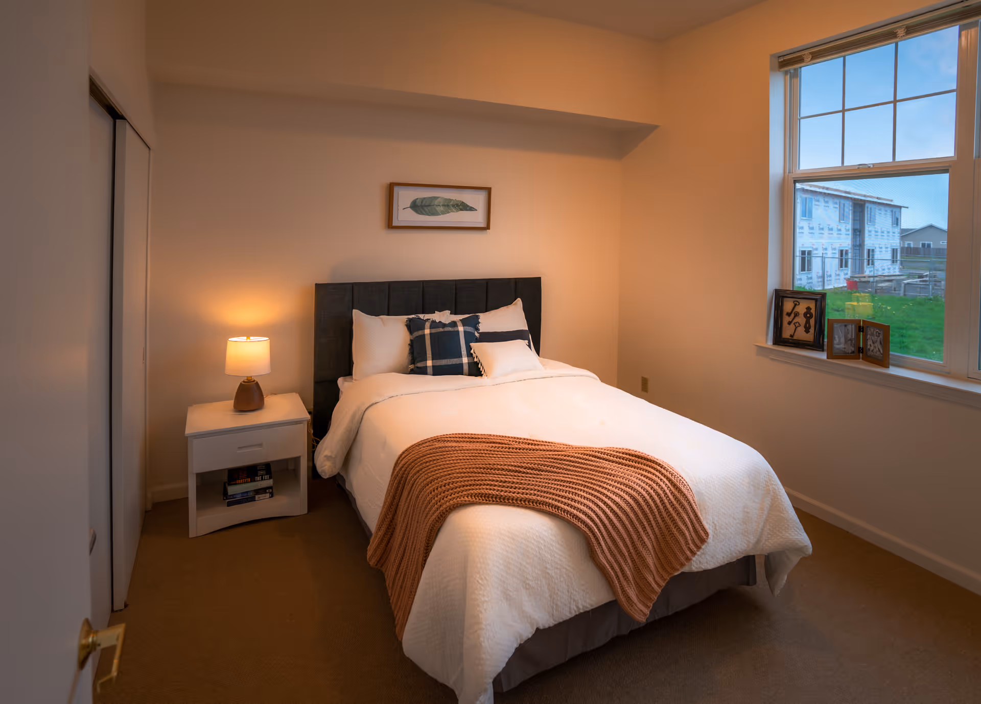 Neatly staged bedroom with a made bed, knitted throw, bedside table and lamp, framed art above the headboard, and a window showing an exterior building.