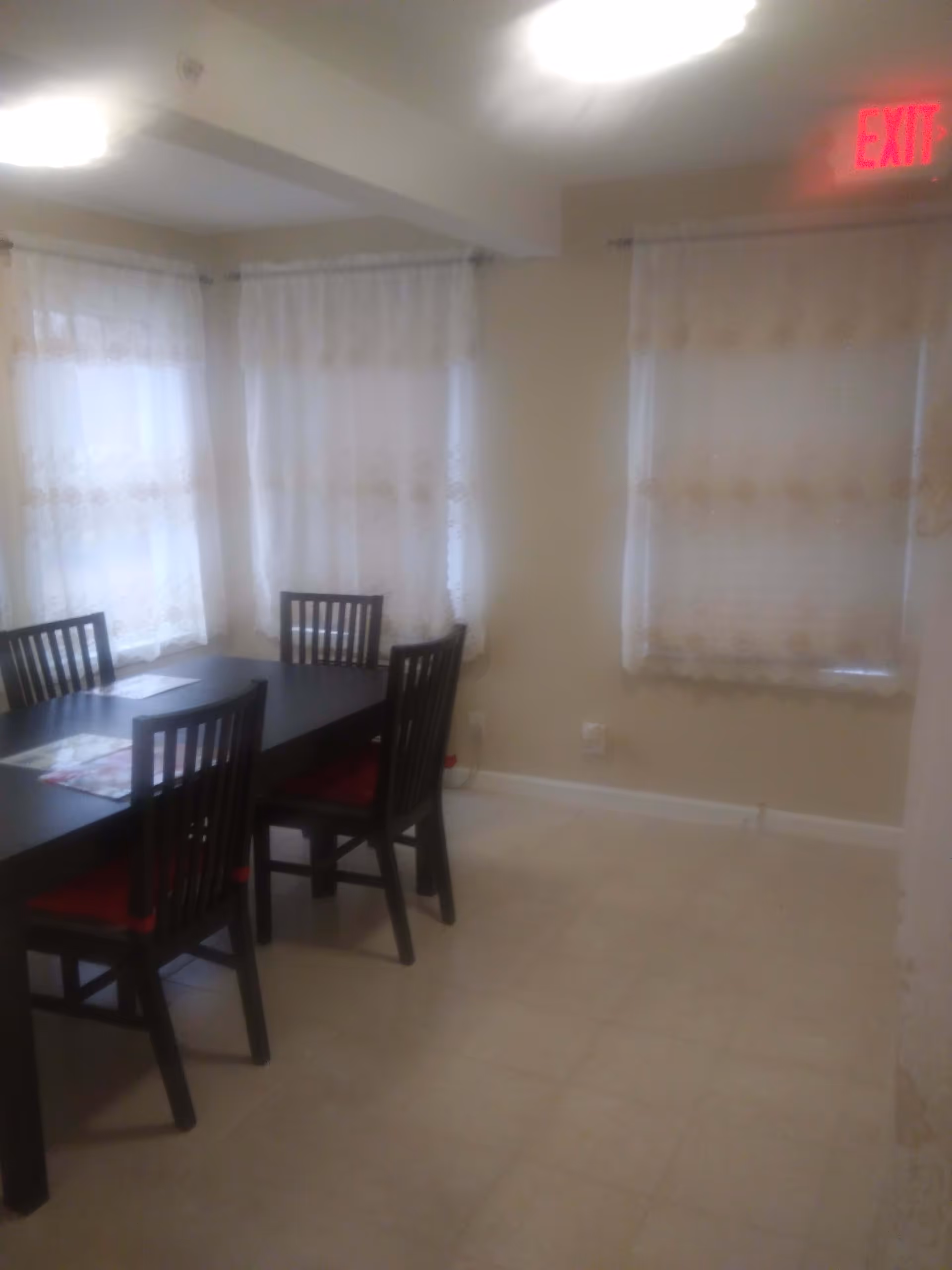 A dining area with a dark rectangular table and four wooden chairs with red cushions. The room has beige walls, tiled floor, and three windows with sheer white curtains. A red exit sign is visible above one window.