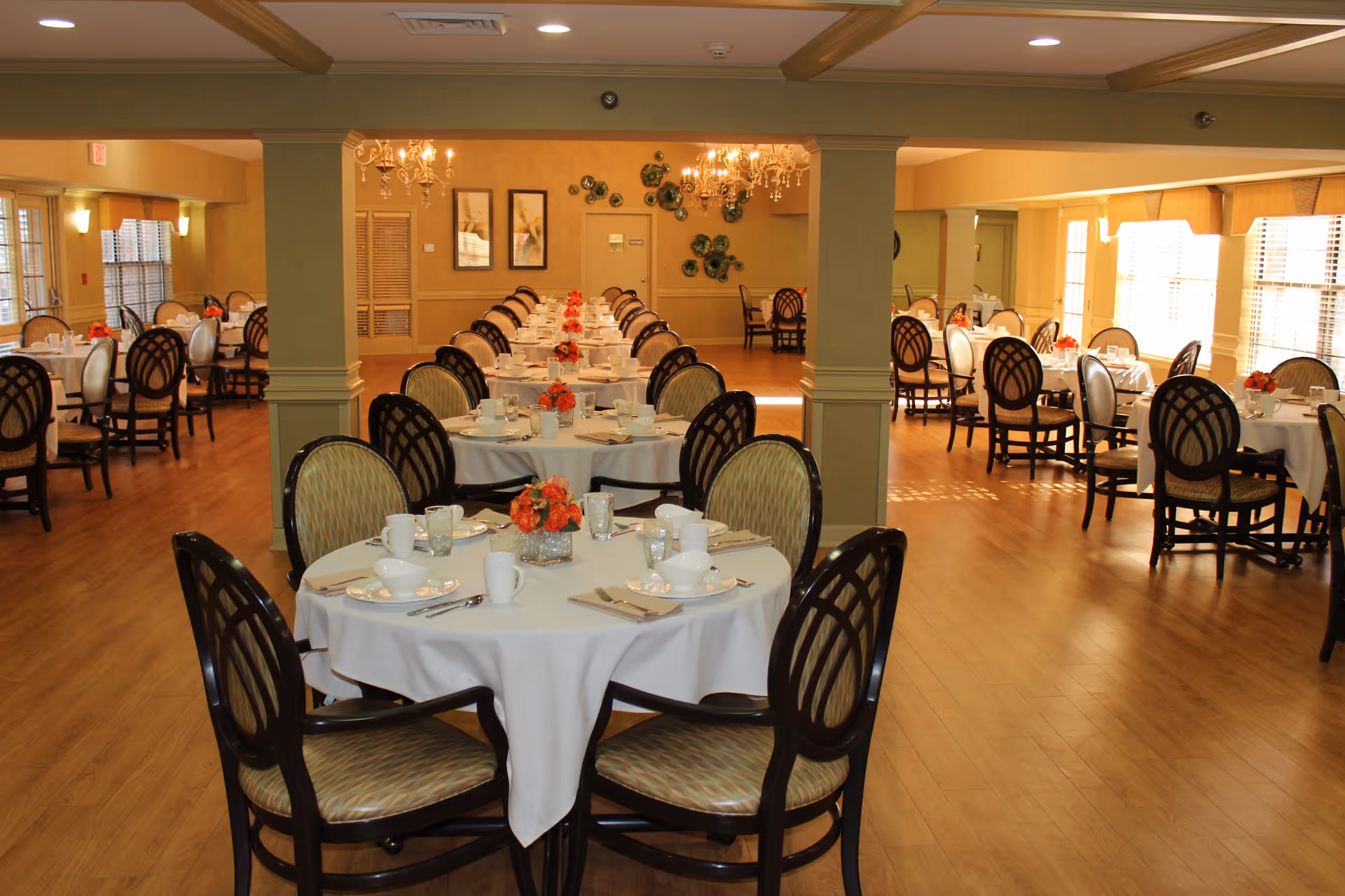 A spacious dining room in a senior living community with multiple round tables covered with white tablecloths, each set with plates, cups, silverware, and small floral centerpieces. The room has wooden flooring, beige walls, and large windows allowing natural light to fill the space.