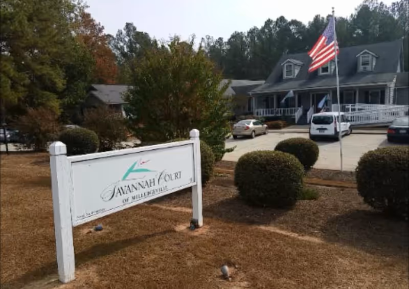 Outdoor view of Savannah Court of Milledgeville facility showing a white sign with the facility name, trimmed bushes, an American flag on a flagpole, a parking area with several cars, and a building with a porch and ramp in the background surrounded by trees.