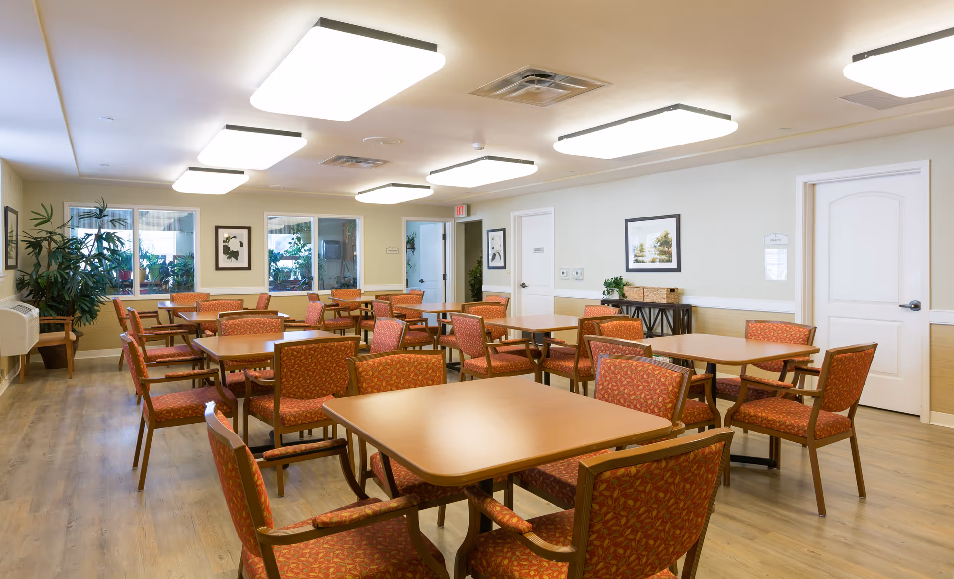 A well-lit room with multiple square wooden tables surrounded by red patterned chairs. The room has light-colored walls, wooden flooring, large ceiling lights, and several framed pictures on the walls. There are also some plants near the windows and a door labeled 'Crafts'.