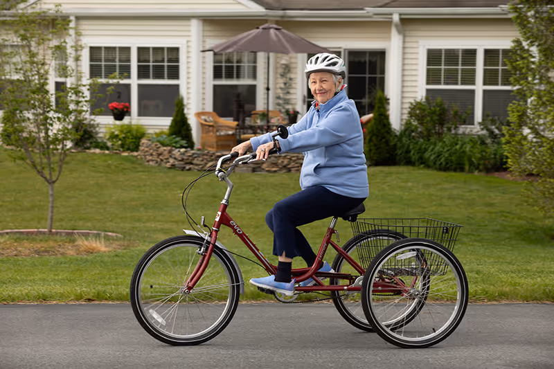 An elderly woman wearing a helmet and blue jacket rides a red three-wheeled bicycle on a paved path in front of a residential building with a patio and greenery.