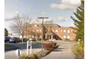 Front exterior of a two-story brick senior care building with a covered entrance, circular driveway, parked cars, and landscaped shrubs and trees.