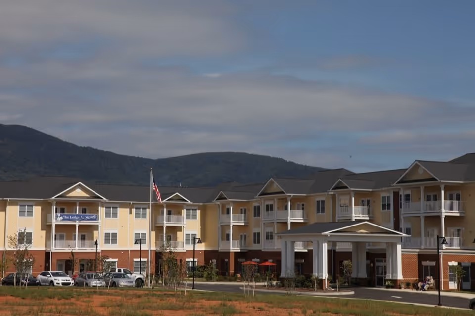 Exterior view of The Lodge at Old Trail, a three-story senior living facility with beige and brick facade, balconies, an entrance canopy, parked cars, and an American flag in front, set against a backdrop of green hills and a partly cloudy sky.