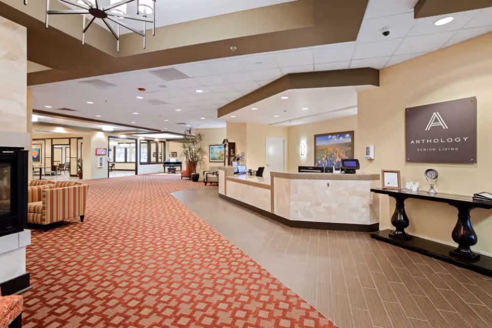 Spacious senior living facility lobby with a reception desk on the right, a patterned red carpet covering the floor, comfortable seating area with striped armchairs on the left, and decorative plants and artwork on the walls.