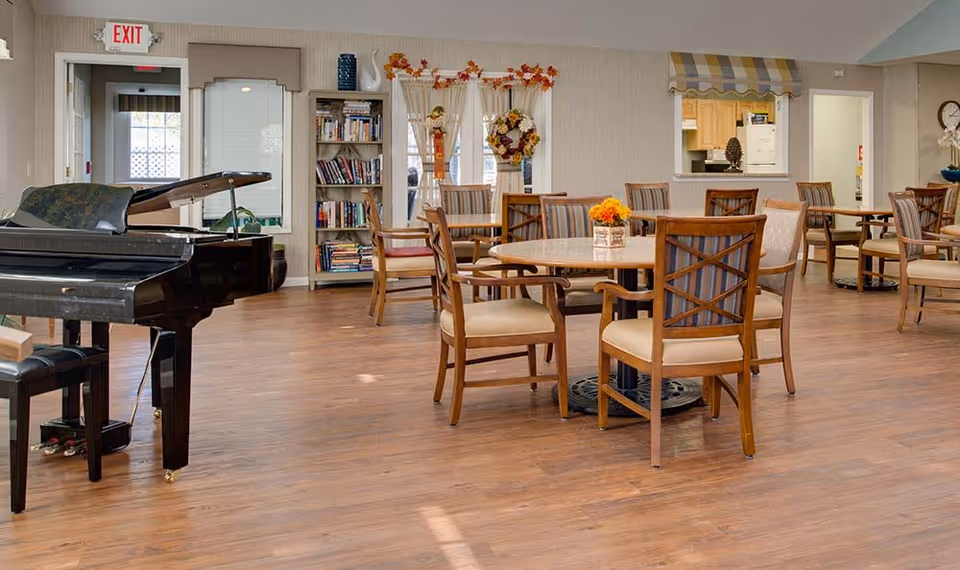 A spacious common area in a senior living facility featuring a black grand piano with a bench, several round wooden tables with cushioned chairs, a bookshelf filled with books, and a kitchen pass-through window. The room has wooden flooring, light-colored walls, and decorative fall-themed wreaths and flowers. An exit sign is visible above a door in the background.
