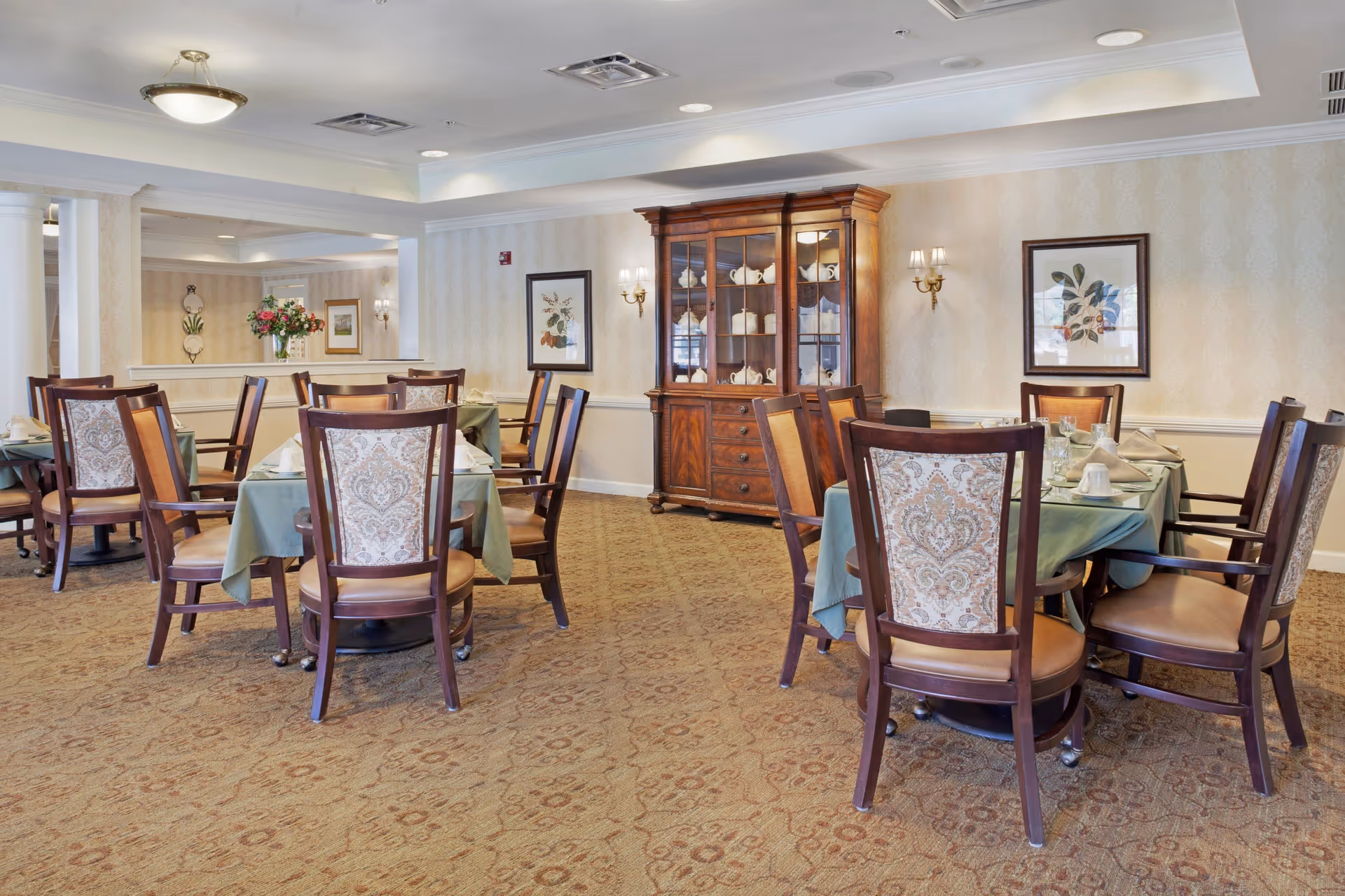 A dining room with several wooden tables covered with green tablecloths and surrounded by wooden chairs with patterned upholstery. A wooden china cabinet filled with white dishware is against the wall, which is decorated with framed botanical prints and wall sconces. The room has a beige patterned carpet and soft lighting from ceiling fixtures.