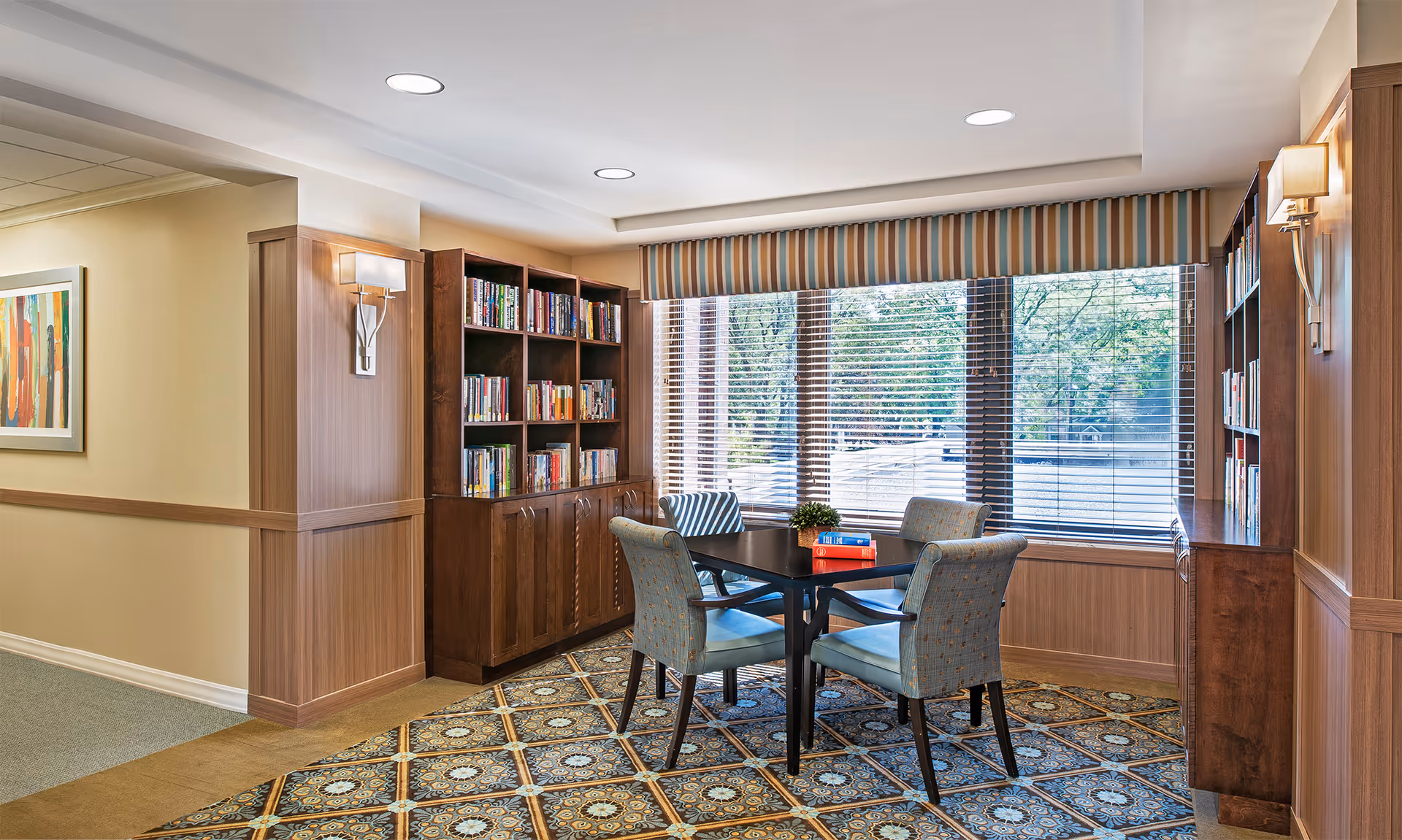 A cozy reading nook in a senior living facility with a square black table surrounded by four upholstered chairs. The area features two wooden bookshelves filled with books, a large window with blinds and a striped valance, patterned carpet, and wall sconces providing soft lighting.