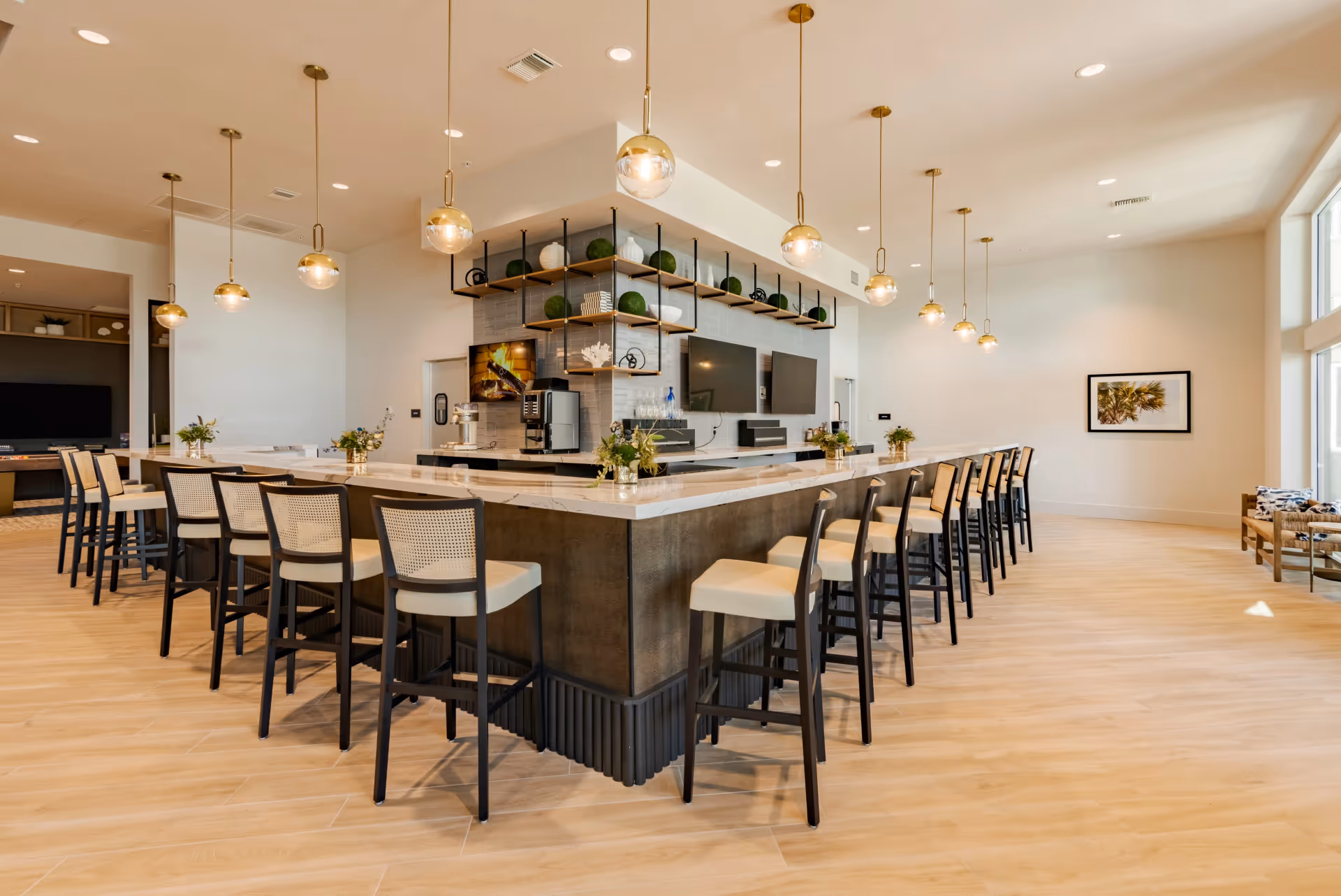 A modern, spacious interior room with a large U-shaped counter surrounded by high chairs with light-colored seats and dark frames. The counter has a white marble top and is decorated with small flower arrangements. Above the counter hang multiple gold pendant lights with round glass bulbs. Behind the counter, there are two large flat-screen TVs mounted on a tiled wall, shelves with decorative items, and a coffee machine. The room has light wood flooring, large windows letting in natural light, and a framed picture on the far wall.