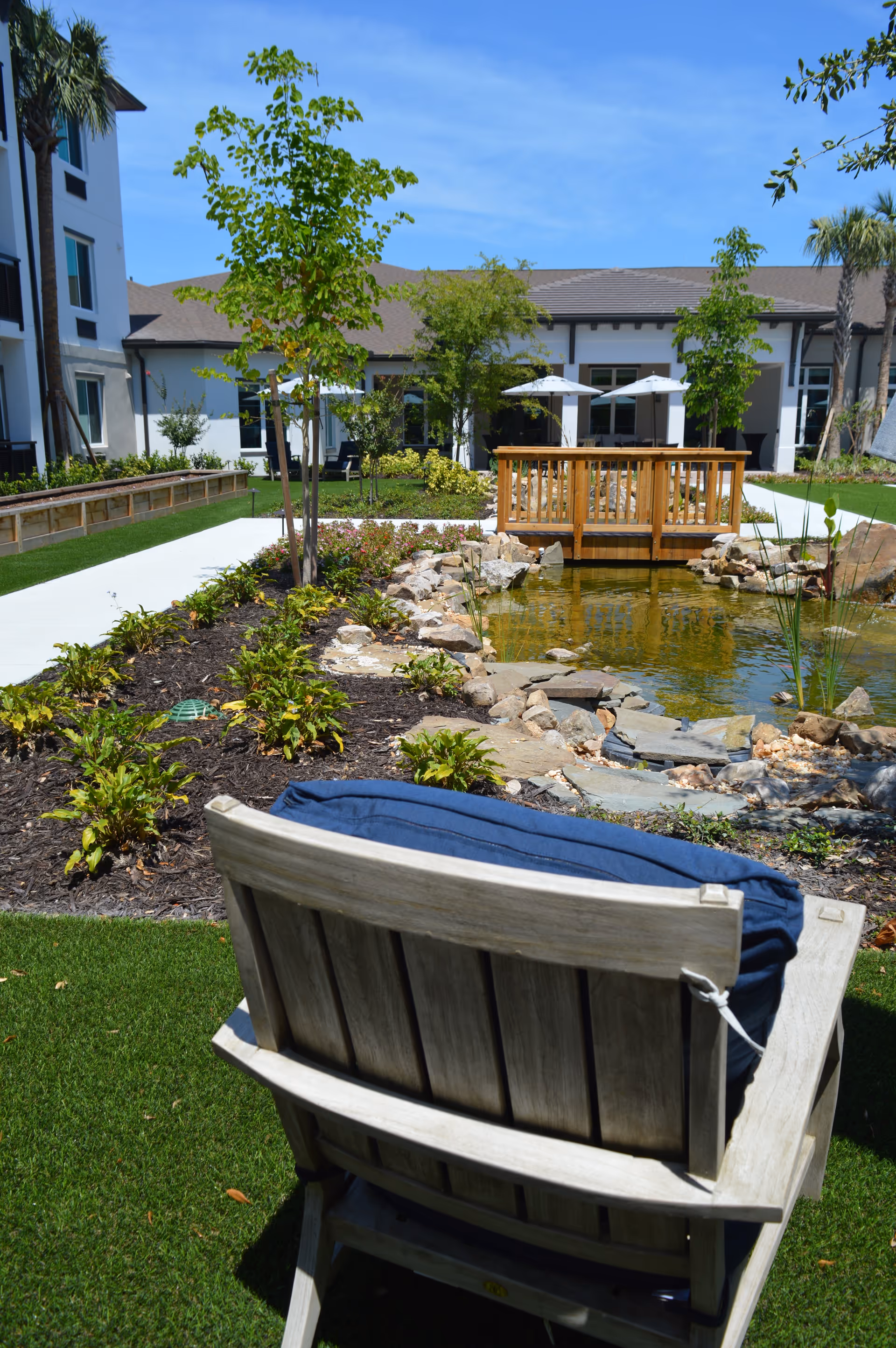 A wooden outdoor chair faces a landscaped pond with a small wooden bridge, patio umbrellas, trees, and the senior living building in the background.
