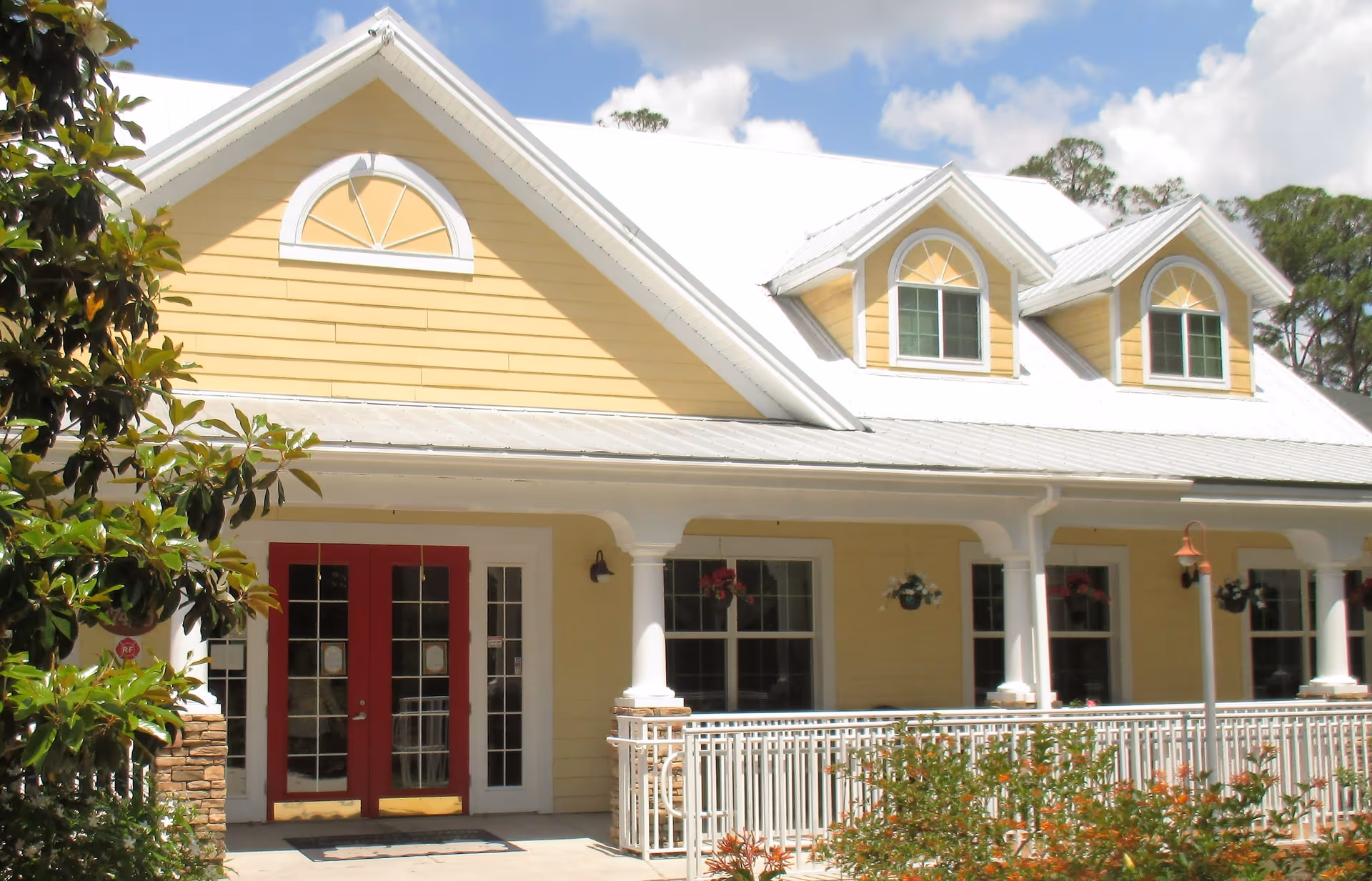 Front exterior view of a yellow building with white trim and a white metal roof. The building has a covered porch with white columns and railings, red double doors, and several windows with decorative arches above them. There are hanging flower pots on the porch and greenery in front of the building under a partly cloudy blue sky.