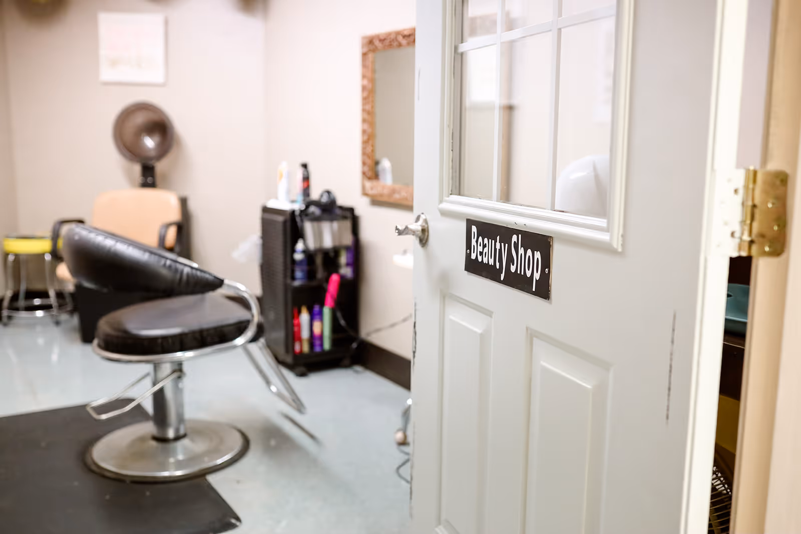 Interior view of a beauty shop with a black salon chair, a hair dryer mounted on the wall, a mirror, and a cart with hair care products. The door to the room is open and has a sign that reads 'Beauty Shop'.