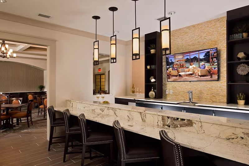 Interior view of a senior living facility bar area with a marble countertop and four black leather bar stools. Three pendant lights hang from the ceiling above the counter. Behind the bar is a wall-mounted flat-screen TV showing a talk show with three people seated. The wall behind the TV has a textured beige tile finish with built-in black shelves holding decorative items and plants. To the left, there is an open doorway leading to a dining area with tables and chairs.
