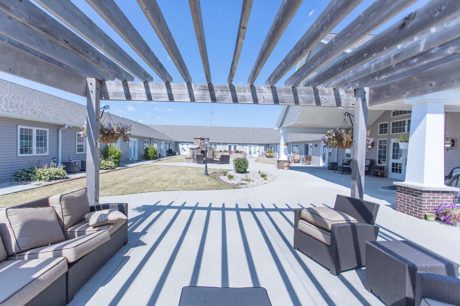 Outdoor seating area with cushioned wicker furniture under a wooden pergola casting striped shadows on the concrete patio. In the background, there are single-story buildings with windows and doors, hanging flower baskets, and a central landscaped area with grass and shrubs under a clear blue sky.