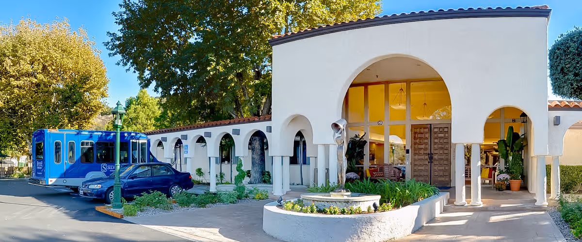 Exterior view of Silverado Calabasas Memory Care Community building with white stucco walls, arched entryways, a small fountain in front, and a blue shuttle bus parked nearby under trees on a sunny day.