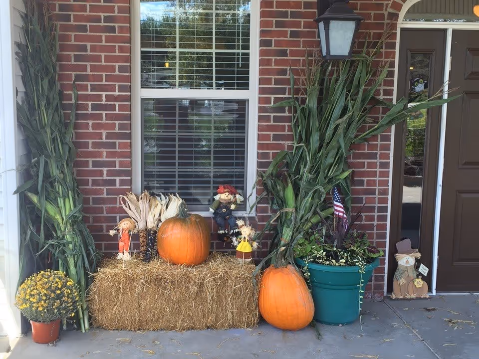 Front porch decorated with autumn harvest items including two large pumpkins, a bale of hay, corn stalks, potted plants, and small scarecrow figurines against a brick wall with a window and a brown door.