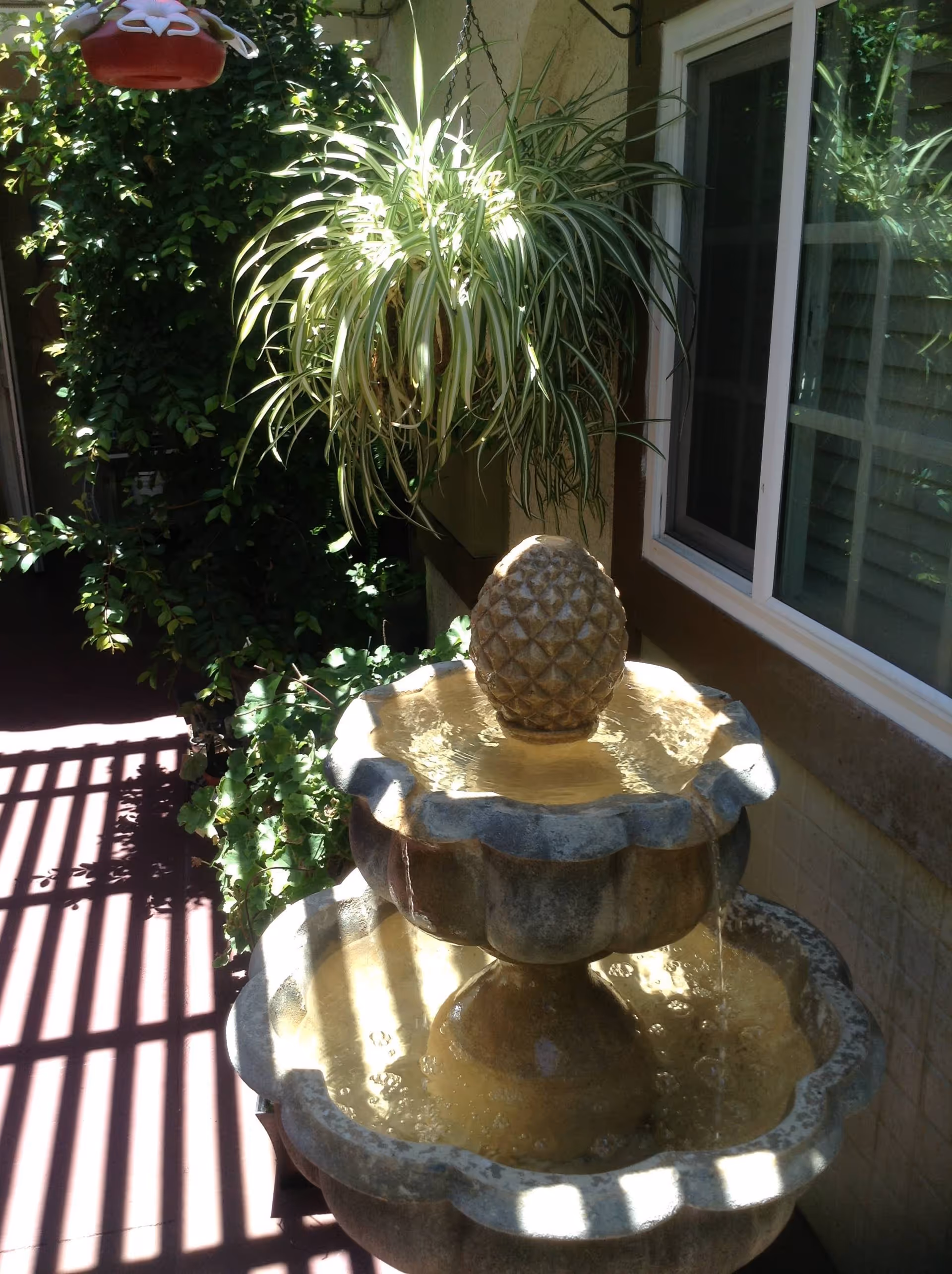 A two-tiered stone water fountain with a pineapple-shaped top piece, surrounded by green plants including a hanging spider plant, located next to a window with sunlight casting shadows through a slatted structure.