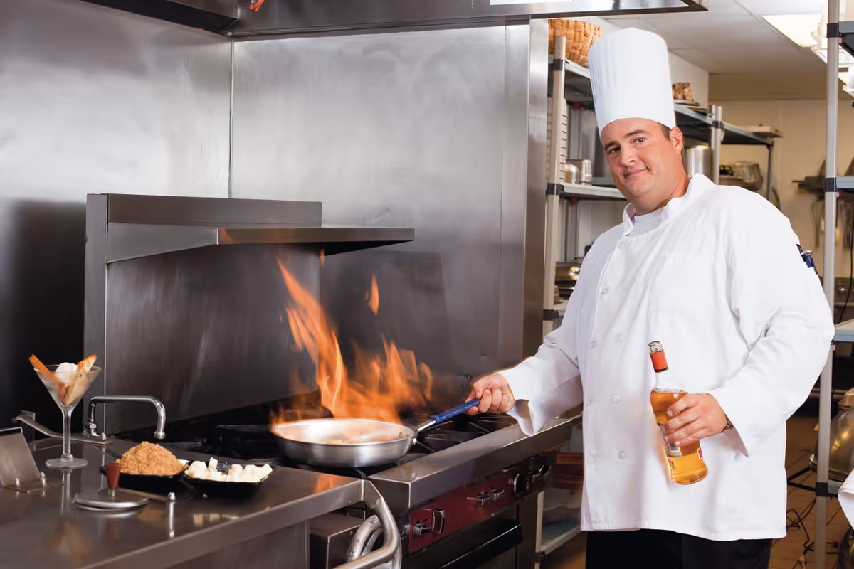 A chef in a white uniform and tall chef hat is cooking with a flaming pan on a stove in a professional kitchen, holding a bottle in one hand and smiling at the camera.