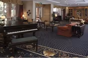 Spacious furnished senior living common room with a grand piano, sofas, tables, and patterned carpet.