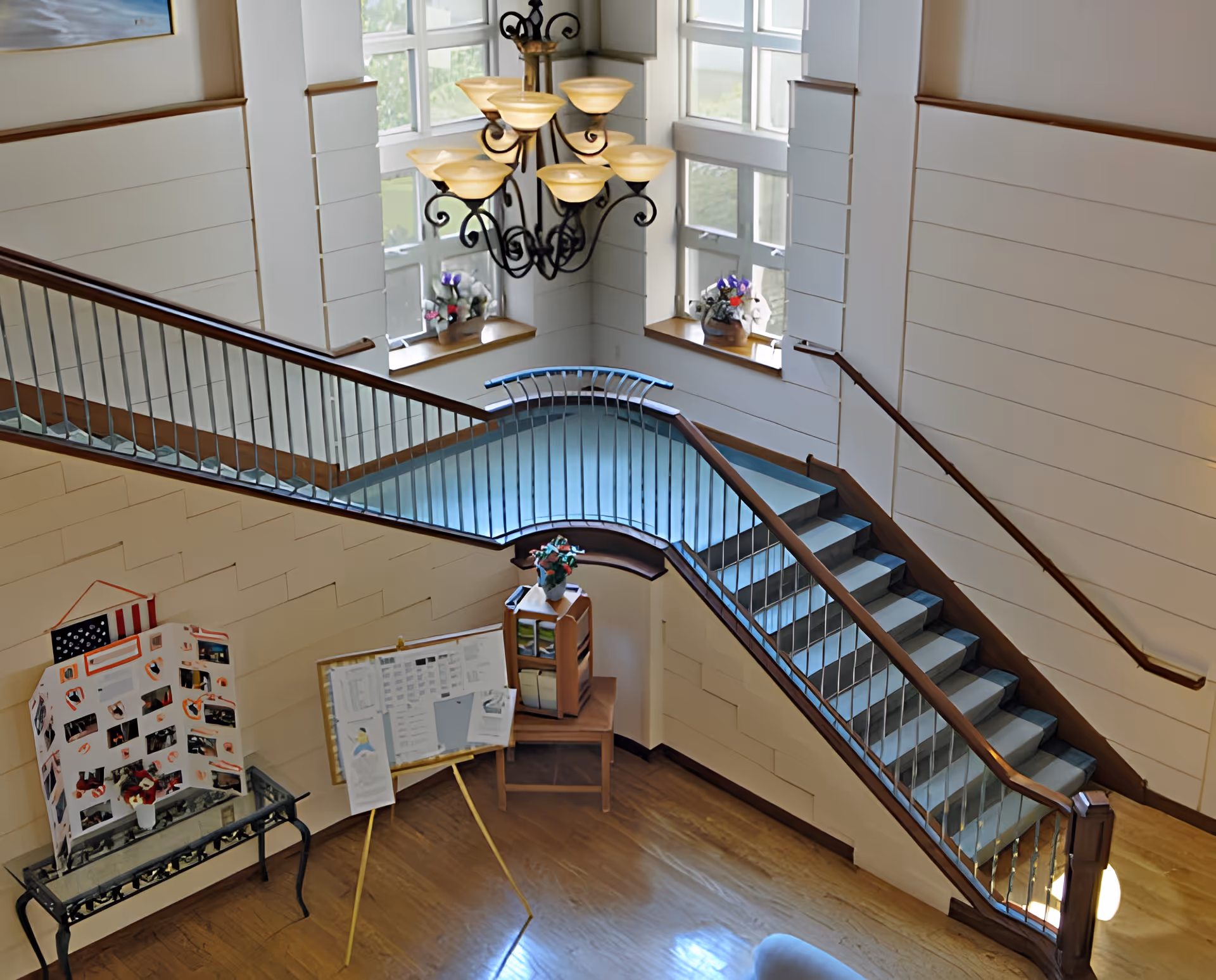 Bright double-height interior stairwell and landing with wooden handrails, chandelier, tall windows and a small display board in the lobby.
