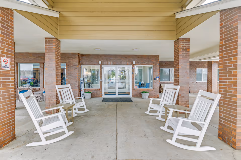 Covered entrance of a senior living facility with white rocking chairs arranged on either side leading to glass double doors.
